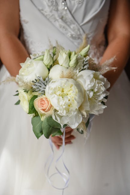 A bridal bouquet featuring a harmonious arrangement of fresh white peonies, roses, and lilies, accented with delicate pale green and gray foliage. The bouquet is gathered into a compact, rounded shape