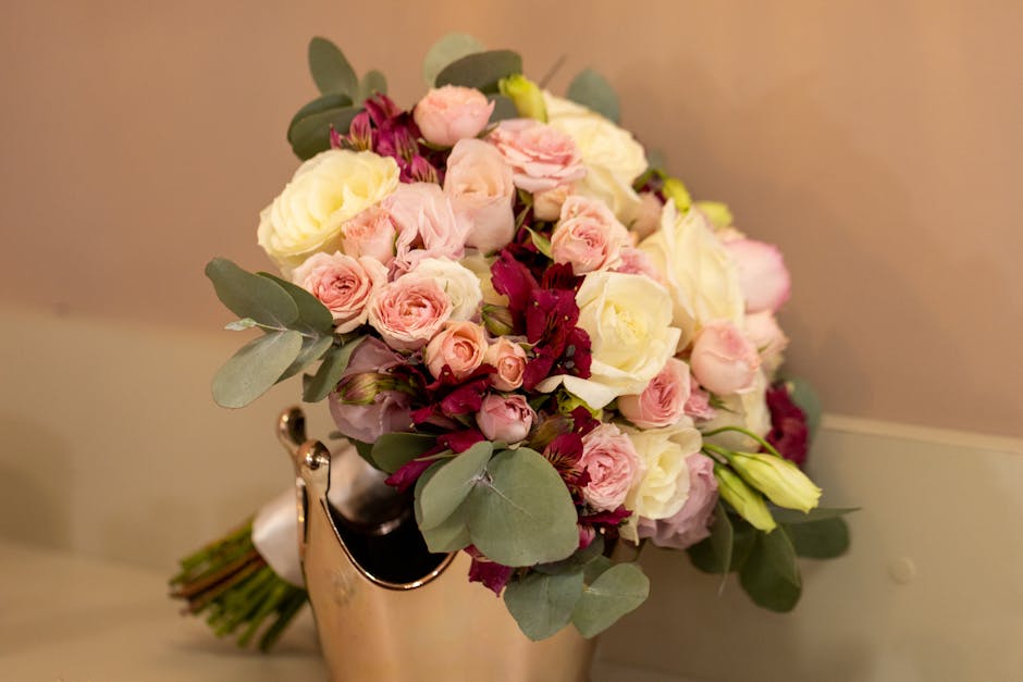 A floral arrangement featuring a bouquet of fresh flowers, predominantly soft pink and creamy white roses, interspersed with dark burgundy blooms and delicate lisianthus. The bouquet includes round, t