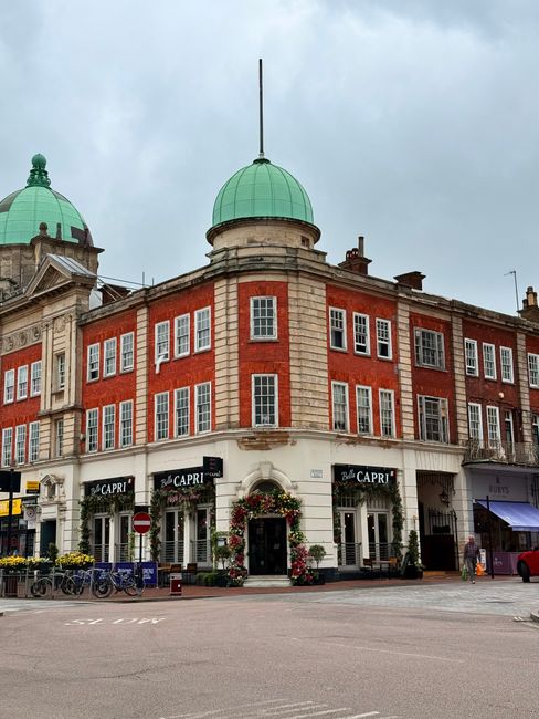The image shows a historic red brick building with white window frames and decorative stone details, topped with two domed green roofs and a central flagpole. The ground floor features large windows w