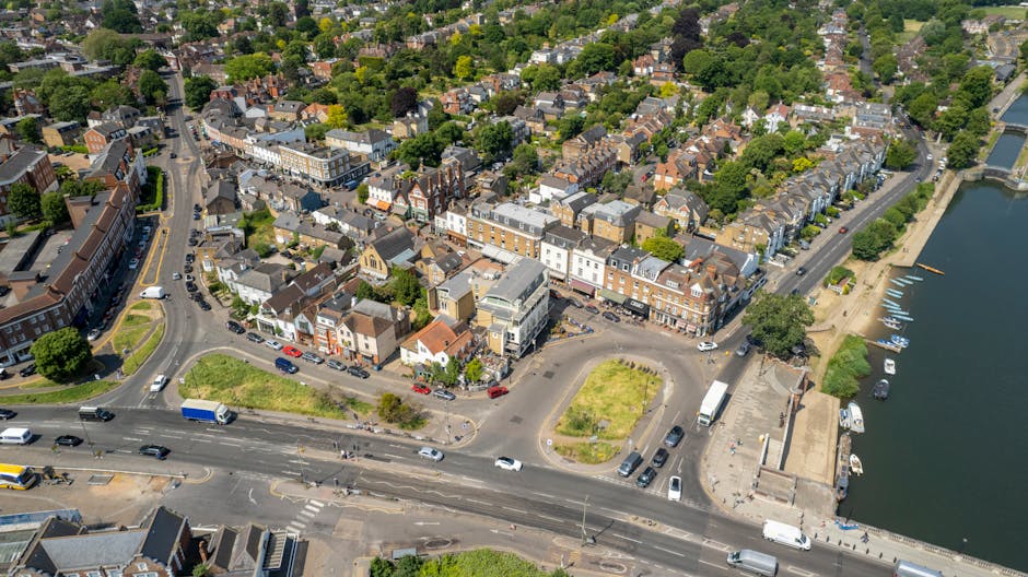 Aerial view of Thames Ditton town centre showing a mix of shops and buildings along the streets, with a river and moored boats on the right side. The scene includes greenery from trees and small parks